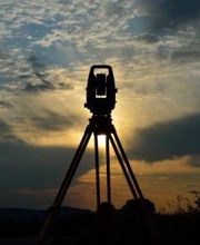 Vermessungsgerät auf Stativ bei Sonnenuntergang, vor bewölktem Himmel.