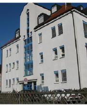 Mehrstöckiges Gebäude mit weißen Wänden und blauen Fensterrahmen, sonniger Himmel.