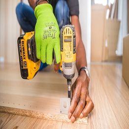 Person in gloves using a power drill on a wooden board indoors.