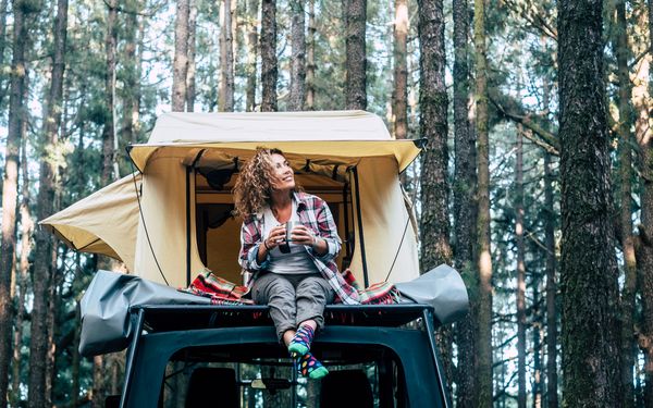 Frau sitz mit Kaffee in der Hand auf dem Dach ihres Autos in ihrem Autodachzelt.