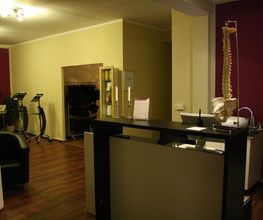 Empfangsbereich mit Schreibtisch, Stühlen, Wirbelsäulenmodell und Fitnessgeräten im Hintergrund.