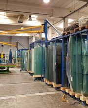 Glasplatten in einer Fabrikhalle, gelbe Kräne und gestapelte Paletten im Hintergrund.