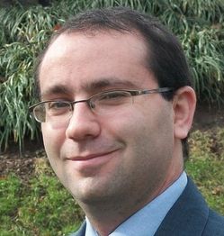 Man with glasses smiling outdoors in a suit, with greenery in the background.