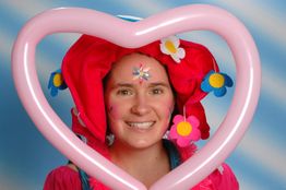 Frau mit Blumenhut lächelt durch ein herzförmiges Luftballon, vor blauem Hintergrund.