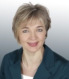 Portrait einer lächelnden Frau mit kurzem blondem Haar und Perlenkette, in einem blauen Blazer.