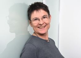 Eine lächelnde Person mit kurzem Haar und Brille lehnt an einer weißen Wand.