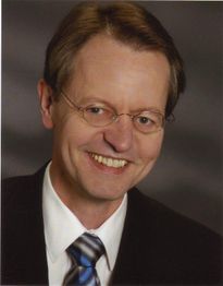 Ein lächelnder Mann in Anzug und Krawatte vor einem grauen Hintergrund.