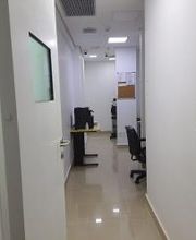 Corredor de escritório com mesas, cadeiras e paredes brancas. Porta aberta em primeiro plano.