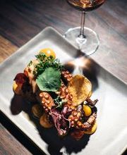 Prato sofisticado com polvo, ervas e legumes coloridos, acompanhado de bebida em taça.