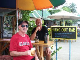 Zwei Männer sitzen vor einer Bar im Freien mit einem Getränk, Sonnenschirme im Hintergrund.