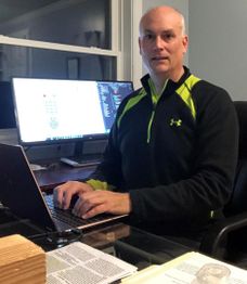 Man sitting at a desk with a laptop, wearing a black and green shirt, looking at the camera.