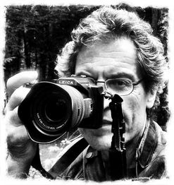 Mann mit Brille hält eine Leica-Kamera vor einem unscharfen Hintergrund. Schwarz-Weiß-Foto.