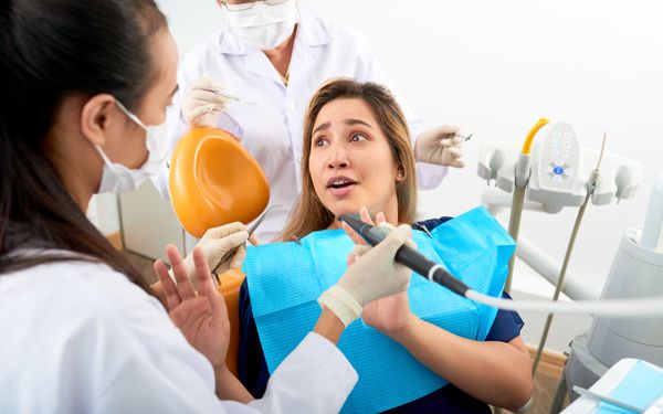 Ängstliche Patientin sitzt im Behandlungsstuhl, während zwei Zahnärztinnen mit Instrumenten eine Untersuchung vorbereiten.