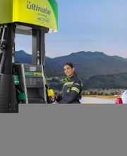 Una mujer en uniforme junto a un surtidor de gasolina con montañas al fondo.