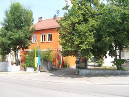 Rotes Gebäude mit Bäumen und flaggen geschmückt, an einer Straße bei sonnigem Wetter.