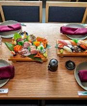 Mesa con sushi, bandejas de sashimi y nigiri, platos, copas, botella de sake y servilletas moradas.