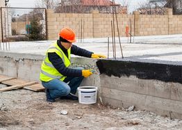Worker applying sealant to a concrete foundation, wearing a safety vest and helmet.