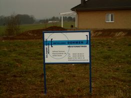 Baustellenschild von Metallbau Dohmen vor einem Haus auf einer grünen Wiese unter grauem Himmel.