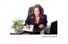 Geschäftsfrau am Schreibtisch mit Laptop und Pflanze, lächelt und schreibt in einen Planer.