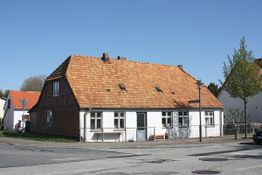 Altes Backsteinhaus mit Satteldach an einer Straßenecke, neben einem Baum unter blauem Himmel.