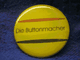 Gelber Button mit der Aufschrift "Die Buttonmacher", zwei diagonale Linien.