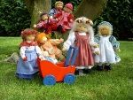 Puppen in traditioneller Kleidung und ein roter Puppenwagen vor einem Baum im Garten.