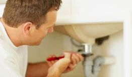 Homem branco reparando encanamento sob pia com chave vermelha.