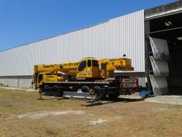 Guindaste amarelo estacionado ao lado de um galpão metálico em um dia ensolarado.