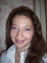 Frau mit langen Haaren lächelt vor einer blumigen Tapete, trägt eine weiße Bluse.