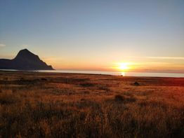 Tramonto su un prato dorato con montagna e mare sullo sfondo, cielo limpido.