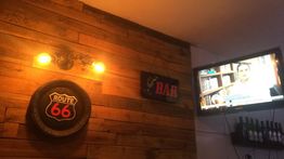 Parede de madeira com placa "Route 66", "BAR" e TV ligada, garrafas na prateleira.