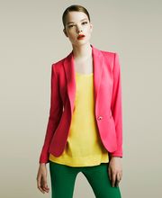 Donna in giacca rosa, top giallo e pantaloni verdi su sfondo grigio chiaro.