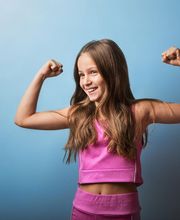 Niña en ropa deportiva rosa sonríe mostrando sus músculos contra un fondo azul.