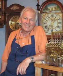 Man in oranje polo en blauwe schort naast antieke klokken.