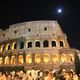 Colosseo illuminato di notte con la luna e folla di persone in movimento.