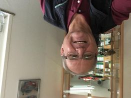 Ein Mann steht lächelnd in einem Büro mit Regalen, Foto ist auf dem Kopf.