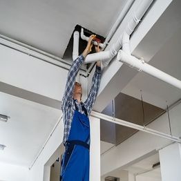 Worker in overalls fixing ceiling pipes in a building.