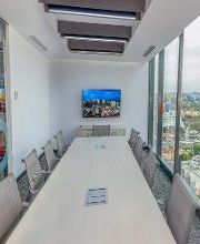 Sala de juntas moderna con mesa larga, ventanas grandes y vista a la ciudad. Pantalla en la pared.