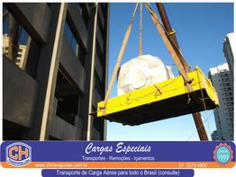 Carga amarela sendo içada por guindaste ao lado de edifício, céu azul ao fundo.