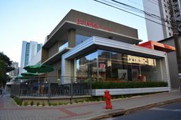 Restaurante de esquina moderno com área externa e guarda-sóis verdes. Edifícios ao fundo.