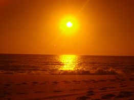 Sonnenuntergang über dem Meer, gelbes Licht reflektiert auf den Wellen, sandiger Strand im Vordergrund.