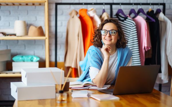 Frau arbeitet mit Laptop und Notizen in einem kleinen Modegeschäft mit Kleidung und Versandkartons.
