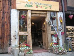 Kleiner, rustikaler Laden mit Holzdekor und Pflanzen vor der Tür, unter dem Schild "Die Wildkräuterey".