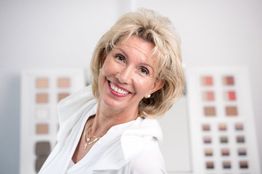 Ältere Frau lächelt vor unscharfem Hintergrund mit Farbkarten, trägt weiße Bluse und Halskette.