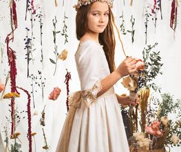 Niña con vestido blanco y corona de flores, rodeada de decoraciones florales colgantes.