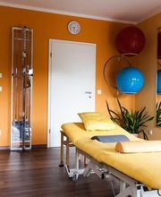 Heller Raum mit Therapieliege, orangenen Wänden, Gymnastikbällen und großen Spiegeln.