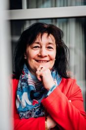 Eine Frau in roter Jacke lächelt, gestützt auf die Hand, vor einem Fenster sitzend.