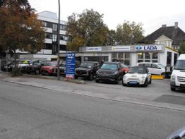 Autos parken vor einem Autohaus mit LADA-Schild, umgeben von Bäumen und anderen Gebäuden.