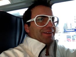 Mann mit weißer Brille in einem Zug, trägt eine weiße Jacke, Hintergrund leicht verschwommen.