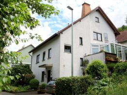 Zweistöckiges, weißes Haus mit Garten und Wintergarten unter blauem Himmel.